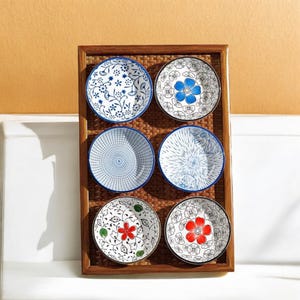 Puede incluir: Seis platos pequeños y redondos de cerámica con diseños florales en azul, rojo y blanco, presentados en una bandeja rectangular de madera. Cada plato presenta un patrón único, incluyendo flores, vides y formas geométricas. Los platos tienen un borde azul o negro.
