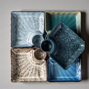 May include: Overhead view of five square ceramic serving dishes in shades of blue, teal, and beige. Each dish has a small, attached circular bowl. The dishes have textured surfaces and are arranged in a square pattern.