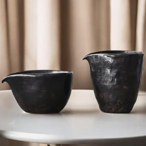 Puede incluir: Dos jarras de cerámica negra con acabado mate se exhiben sobre una superficie blanca. Las jarras tienen un pico para verter. La jarra de la izquierda es más ancha y corta que la jarra más alta de la derecha. El fondo es de color neutro.