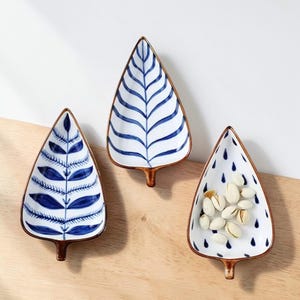 Puede incluir: Tres platos de cerámica con forma de hoja con diseños azules y blancos. Un plato contiene pistachos. Los platos tienen borde y tallo marrón. Los diseños incluyen vetas de hojas, gotas y diseños abstractos.