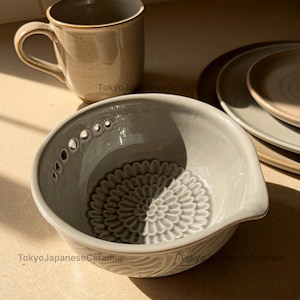 Può includere: Una ciotola in ceramica grigio chiaro con un motivo floreale testurizzato alla base e una fila di piccoli fori lungo il bordo interno, accompagnata da una tazza maculata abbinata e due piatti impilati, tutti con il testo TokyoJapaneseCeramic.