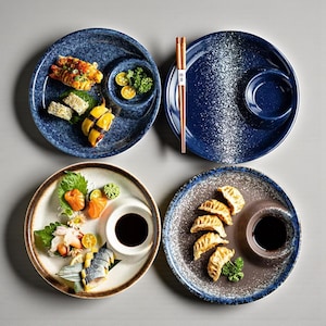 Puede incluir: Cuatro platos de sushi de cerámica con varios sushi y salsas. Dos platos son azules con motas blancas, uno es marrón con borde azul y otro es blanco con borde marrón. Palillos chinos descansan sobre un plato.