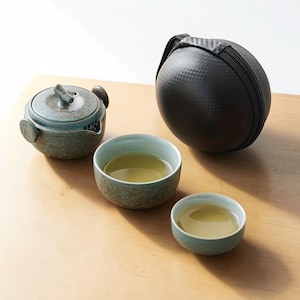 Puede incluir: Un juego de té turquesa con una tetera pequeña y dos tazas, todas llenas de té. También hay un estuche de transporte negro y redondo con cremallera. El juego está sobre una superficie de madera clara.