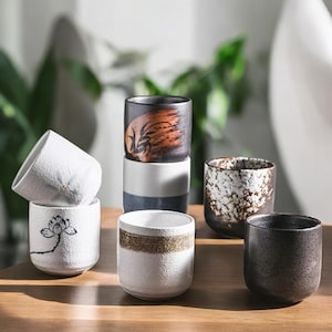 Könnte beinhalten: Eine Sammlung von Keramikbechern mit einzigartigen Designs. Die Becher variieren in der Farbe, darunter Weiß, Schwarz und Braun, und weisen strukturierte Oberflächen und dekorative Muster auf. Einige Becher haben florale Motive oder goldglitzernde Bänder.