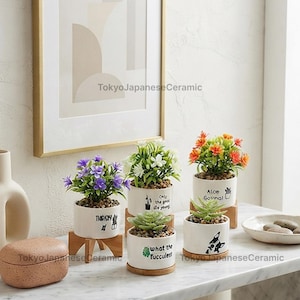 Puede incluir: Cinco pequeñas macetas cilíndricas de cerámica blanca sobre soportes de madera, cada una con una planta artificial diferente. Las macetas presentan texto e ilustraciones en negro, incluyendo frases como 'Only the good die young'.