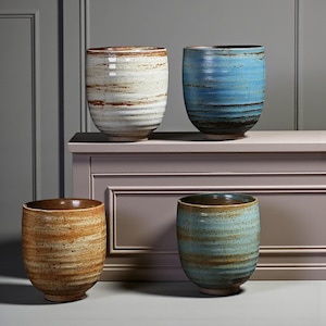 May include: Four ceramic tumblers in white, blue, brown, and tan. Each has a textured, horizontal stripe design. The tumblers are displayed on a light purple surface. The handmade cups showcase a variety of colours and textures.
