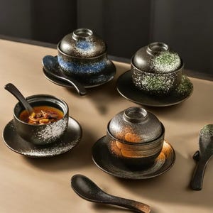 Può includere: Un set di quattro ciotole per zuppa in ceramica nera con coperchi, piattini e cucchiai. Le ciotole presentano un design maculato bianco e colorato, con accenti blu, verdi e arancioni. Una ciotola contiene zuppa.