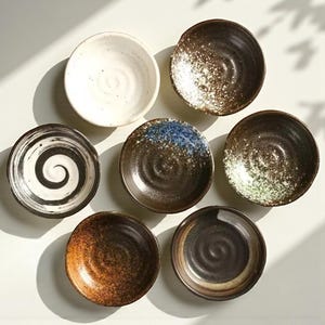 Puede incluir: Colección de siete pequeños platos redondos de cerámica. Los platos presentan una variedad de diseños, incluyendo patrones moteados, una espiral en blanco y negro y colores lisos. La paleta de colores incluye blanco, marrón y azul.