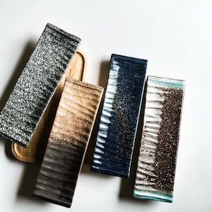 May include: An overhead shot of five rectangular ceramic serving dishes. The dishes have a speckled or gradient design in shades of black, brown, and blue. One dish rests on a wooden tray. The dishes are arranged on a white surface.