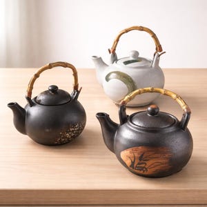 Puede incluir: Tres teteras de cerámica con asas de bambú. Dos son negras con diseños decorativos, y una es blanca con un diseño verde y marrón. Las teteras están sobre una superficie de madera clara.