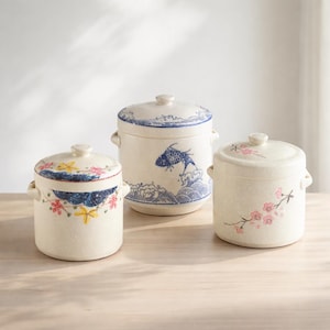 Puede incluir: Tres tarros de cerámica con tapas, cada uno con diseños únicos pintados a mano. Los tarros son de color blanco roto con motivos florales y de peces azules y rosas. Cada tarro tiene un asa pequeña a cada lado y una tapa a juego con un pomo.