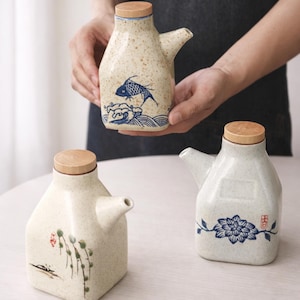 Puede incluir: Tres dispensadores de aceite de cerámica con tapas de madera. Uno presenta un diseño de pez azul, otro un patrón floral y el tercero un diseño de planta. Los dispensadores son de color beige con una textura moteada.