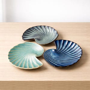 Könnte beinhalten: Drei keramische Muschelschalen in Blau- und Türkistönen. Jede Schale hat ein Spiralmuster mit dunklem Rand. Die Schalen sind auf einer hellen Holzoberfläche angeordnet.
