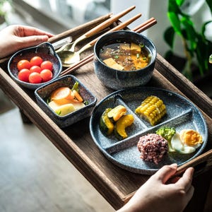 Puede incluir: Una bandeja de madera presenta una comida japonesa. Incluye un cuenco de sopa, un plato dividido con maíz, verduras y arroz, y pequeños cuencos con tomates y otros acompañamientos. También hay palillos y cucharas.