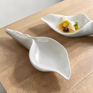 May include: Two white ceramic serving dishes shaped like calla lilies. One dish contains food, including a lime slice. The dishes are on a light-colored wooden surface. The dishes have a smooth, glossy finish.