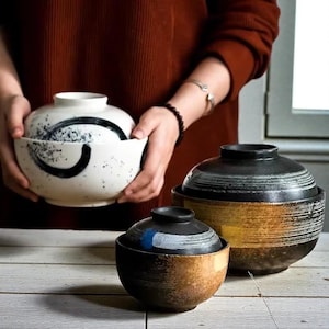 以下が含まれることがあります： 黒い抽象的なデザインの白いセラミックボウルを持つ人。ふた付きの他の2つのセラミックボウルが木製の表面にあります。1つのボウルは黒と金で、もう1つは黒いふた、青と白のアクセントが付いた茶色です。