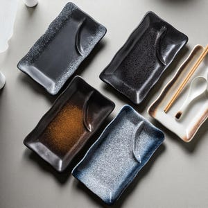 Puede incluir: Colección de platos rectangulares de cerámica en varios colores, incluyendo negro, azul y marrón. Cada plato tiene un acabado moteado o texturizado único y una hendidura curva. Palillos y una cuchara descansan sobre un plato blanco.