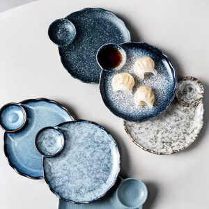 May include: A selection of speckled ceramic plates in blue and white. Each plate has a small attached dipping bowl. Some plates hold dumplings and sauce. The plates have a wavy edge and a rustic aesthetic.