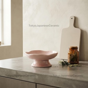 Puede incluir: Un cuenco de pedestal de cerámica rosa claro con borde ondulado, una tabla de cortar de madera clara y un pequeño frasco de vidrio ámbar lleno de hierbas secas. El texto TokyoJapaneseCeramic es visible en la imagen.