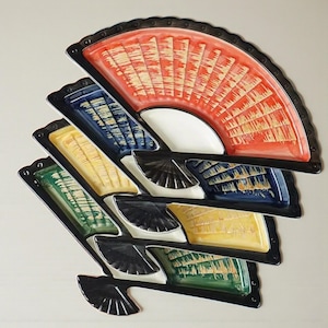 May include: A collection of ceramic fan-shaped serving dishes in various colours. The dishes are red, blue, yellow, and green, with black rims. The fan designs feature textured patterns.