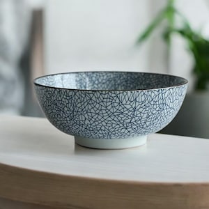 Op de afbeelding: Een keramische kom met een witte basis en een blauw-wit craquelé patroon. De kom heeft een zwarte rand en staat op een lichtgekleurd oppervlak. De kom is leeg en klaar om te worden gevuld.