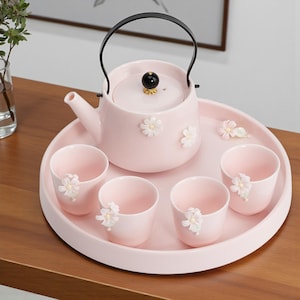 以下が含まれることがあります： ピンク色の陶器のティーセット。ティーポット、カップ4つ、トレイが含まれています。 ティーポットは黒いハンドルと蓋に黒と金のノブが付いています。 各ピースは白い花のアクセントで装飾されています。