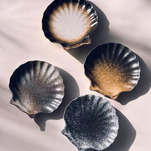 Könnte beinhalten: Vier keramische Muschelschalen in verschiedenen Schwarz-, Weiß- und Goldtönen. Jede Schale hat eine strukturierte, gesprenkelte Oberfläche und einen gewellten Rand. Die Schalen sind auf einer hellrosa Oberfläche angeordnet.