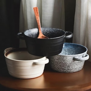 May include: Three ceramic bowls in black, white, and speckled blue-gray, each with two handles. A wooden spoon rests in the black bowl. The bowls are on a brown surface, with a neutral background.