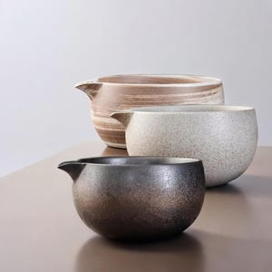 Pode incluir: Três tigelas de cerâmica em vários tamanhos e cores. A tigela maior é marrom claro com listras horizontais. A tigela do meio é esbranquiçada salpicada. A tigela menor é cinza escuro com brilho metálico. Cada tigela tem um bico.