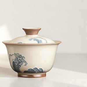 May include: Cream-colored ceramic tea bowl with a matching lid. The bowl and lid feature a blue and gray wave pattern. The rim and the lid's handle are brown. The bowl is designed for tea ceremonies.