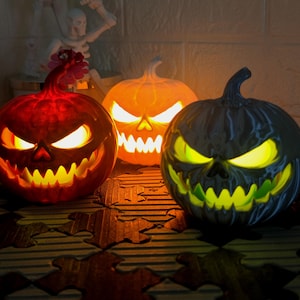 Puede incluir: Tres decoraciones de calabazas de Halloween iluminadas. Las calabazas están talladas con caras amenazantes, con ojos brillantes y dientes de sierra. Las calabazas son en tonos de rojo, naranja y negro. Una figura de esqueleto está en el fondo.