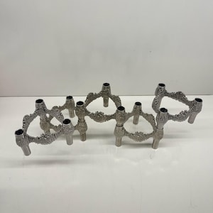 Op de afbeelding: Een set van drie zilverkleurige metalen kandelaars. Elke houder heeft een textuur, organisch ontwerp met meerdere kaarshouders. De houders zijn in een rij gerangschikt, wat hun decoratieve en functionele aspecten benadrukt. De achtergrond is wit.