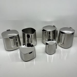 May include: A collection of six stainless steel serving items, including a creamer, sugar bowl, and lidded containers. The pieces have a polished, reflective finish and a modern, minimalist design. The set is likely for serving coffee or tea.