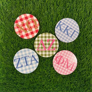 May include: Five circular buttons featuring gingham patterns and Greek letters. One is red and white with gold letters XΩ, one blue and white with blue letters KKT, one green and white with pink letters ΔZ, one light blue and white with blue letters ZTA, and one pink and white with pink letters ΦM.
