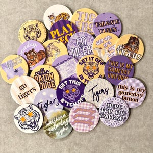 May include: A collection of round pinback buttons featuring various designs in purple, gold, and white. The buttons display tiger illustrations, star-shaped eyes, and text including "Go Tigers," "Tailgate Queen," "Gameday," and "Louisiana Saturday Night."