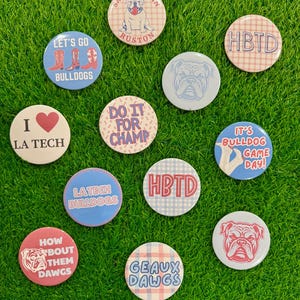 May include: A collection of round buttons with various designs and text, including bulldog graphics and phrases like "Let's Go Bulldogs," "Saturday in Ruston," and "It's Bulldog Game Day!" The buttons are arranged on a green, grassy surface.