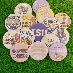 May include: A collection of round buttons with various designs and text, including tiger illustrations and phrases like "Calling Baton Rouge" and "Geaux Tigers." The buttons feature purple, gold, and white colors.