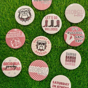 May include: A collection of white and red Georgia Bulldogs themed buttons. The buttons feature various designs, including the team logo, text, and graphics. The buttons are arranged on a bed of green grass.