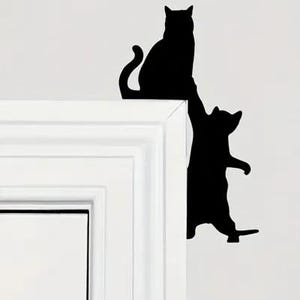 May include: A black wall decal featuring two cat silhouettes. One cat sits on the top edge of a white door frame, while the second cat is positioned vertically along the side of the frame, appearing to climb or reach up toward the first cat.