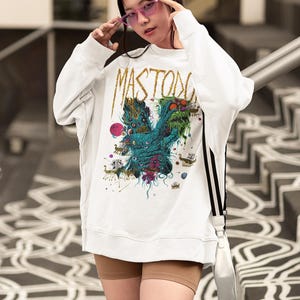 May include: White sweatshirt with a colorful graphic design featuring the word "MASTODON" in gold lettering. The graphic includes a teal monster with multiple eyes and other fantastical elements. The person is wearing pink sunglasses.