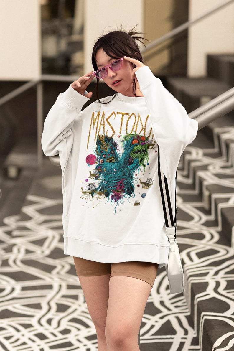 May include: White sweatshirt with a colorful graphic design featuring the word "MASTODON" in gold lettering. The graphic includes a teal monster with multiple eyes and other fantastical elements. The person is wearing pink sunglasses.