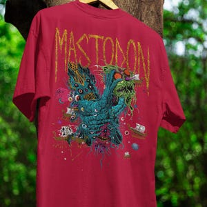 May include: Red t-shirt featuring a teal and green monster graphic with the band name "Mastodon" in gold lettering. The design includes multiple eyes, ships, and a variety of colors. The shirt is hanging on a wooden hanger.