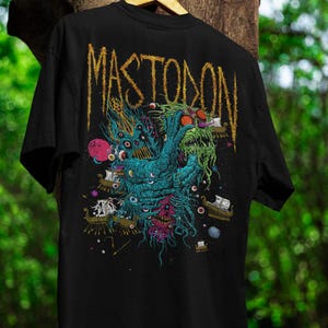 May include: Black t-shirt featuring a detailed, colorful graphic of a creature with multiple eyes, tentacles, and ships. The word "MASTODON" is printed in a distressed gold font across the top. The shirt is hanging on a wooden hanger.