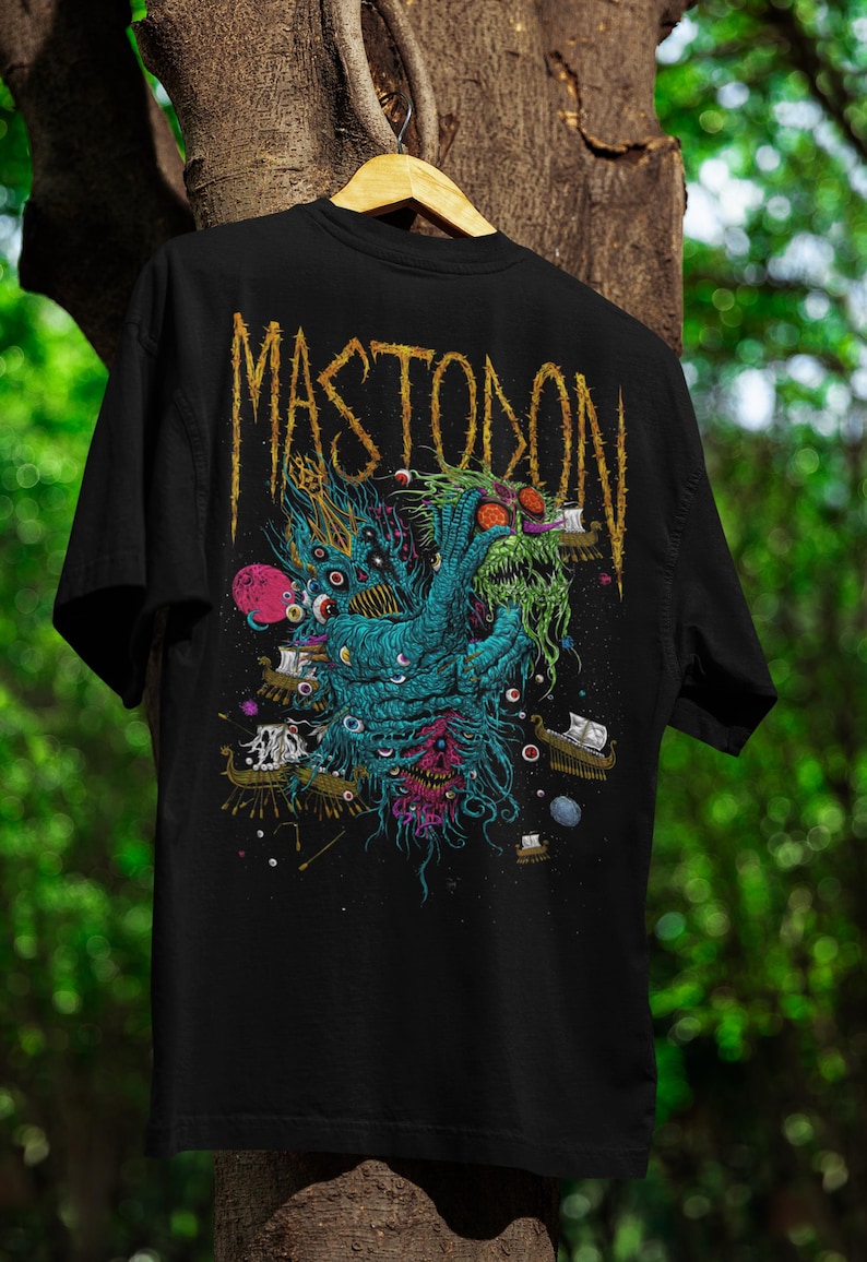 May include: Black t-shirt featuring a detailed, colorful graphic of a creature with multiple eyes, tentacles, and ships. The word "MASTODON" is printed in a distressed gold font across the top. The shirt is hanging on a wooden hanger.