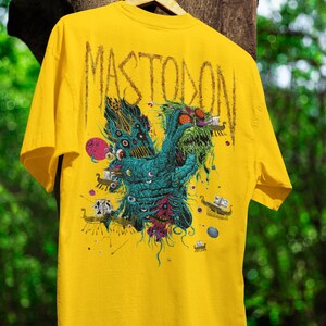 May include: Yellow t-shirt featuring the band name "MASTODON" across the upper back. The front graphic is a detailed, colorful illustration of a creature with multiple eyes and tentacles. The design includes shades of teal, red, and purple.