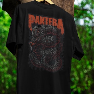 Pode incluir: Camiseta preta com um desenho de cobra enrolada em cinza e vermelho, e o nome da banda "PANTERA" em letras laranja. O gráfico está nas costas, criando um estilo rock and roll.