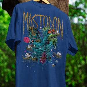 May include: Blue t-shirt with a graphic design featuring the word "MASTODON" in gold lettering. The design includes a monster with multiple eyes, ships, and a red planet. The shirt is hanging on a wooden hanger.