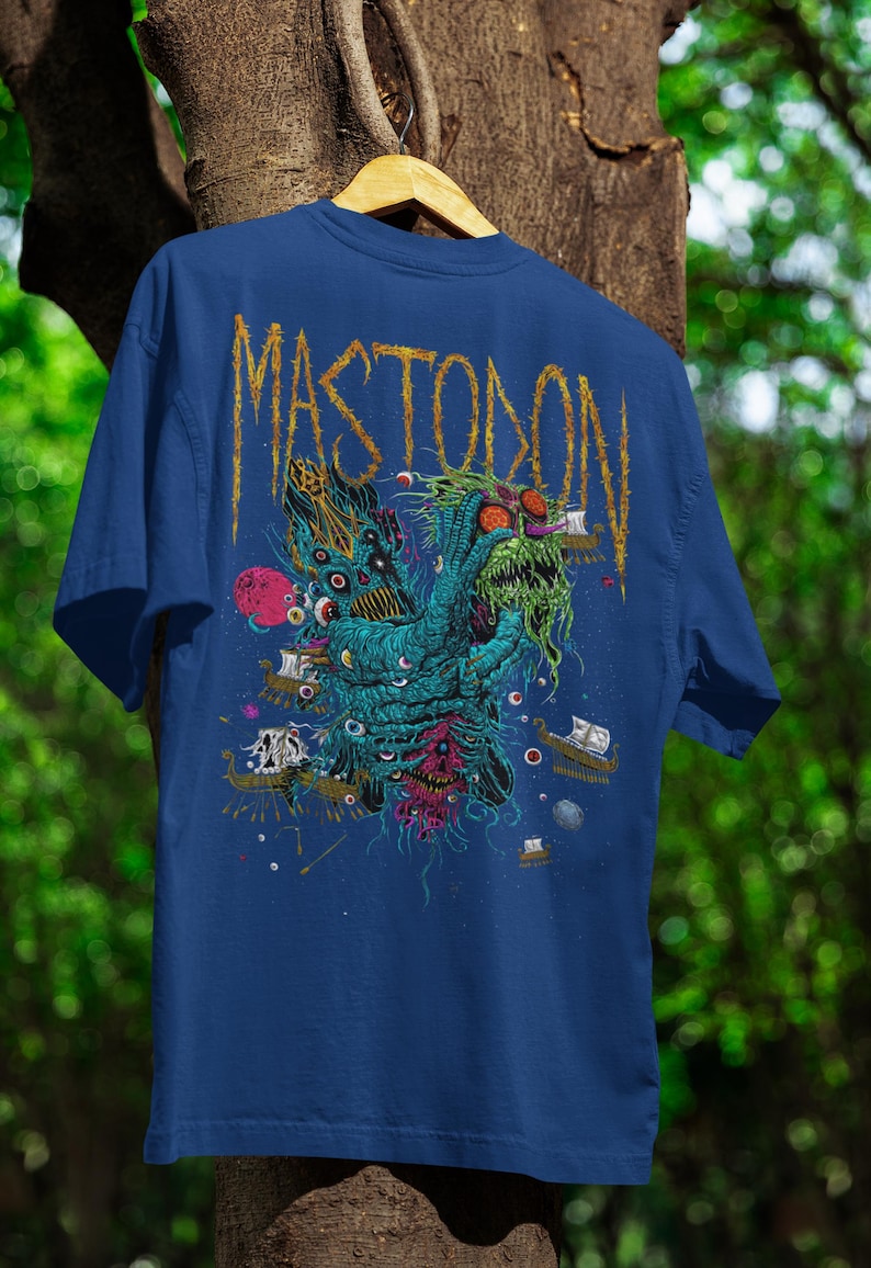 May include: Blue t-shirt with a graphic design featuring the word "MASTODON" in gold lettering. The design includes a monster with multiple eyes, ships, and a red planet. The shirt is hanging on a wooden hanger.