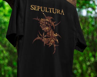 Puede incluir: Camiseta negra con el nombre de la banda "SEPULTURA" en letras doradas en la parte superior. La parte posterior de la camiseta muestra un diseño gráfico detallado en marrón y dorado. El diseño parece ser una criatura abstracta estilizada.