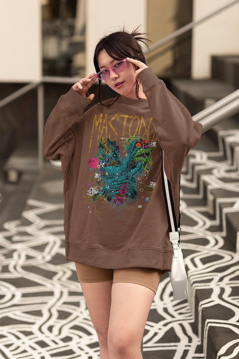 May include: Brown sweatshirt with the word "MASTODON" in gold lettering. The graphic on the front features a colorful monster design with multiple eyes and tentacles. The sweatshirt is worn with tan shorts.